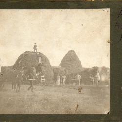 Photograph of hay stacks with people and a team of horses with a wagon