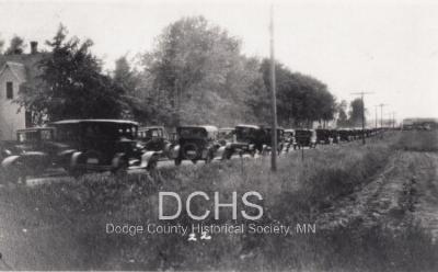 Black and white photograph of many cars lined up on road to see tornado damage