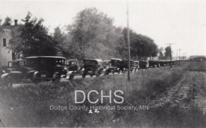 Black and white photograph of many cars lined up on road to see tornado damage